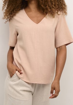 Femme portant un haut col en V à manches courtes rose poudré et un pantalon crème texturé, avec une main dans la poche et des cheveux bouclés visibles.