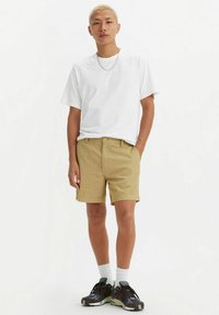 Cămașa albă din bumbac este asortată cu pantaloni scurți kaki. Modelul poartă adidași cu model negru și albastru, împreună cu șosete albe până la glezne. O ținută simplă și lejeră.