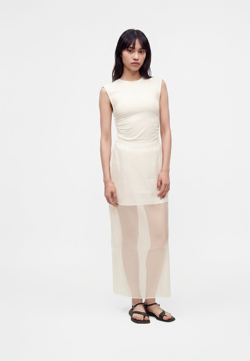 Jeune femme aux cheveux noirs lisses portant une robe blanche sans manches avec une jupe transparente et des sandales noires, debout sur un fond blanc.