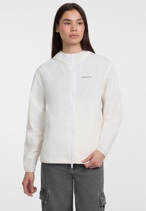 Giacca leggera di colore crema con cappuccio, zip anteriore e dettaglio del logo sul petto; presenta polsini elasticizzati e una vestibilità rilassata.