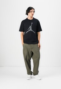 Sort T-shirt med et stort gråt Jumpman-logo, parret med løse olivenfarvede cargo-bukser og hvide sneakers. En simpel, afslappet sportsklædning.
