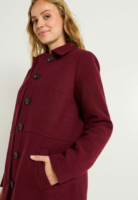 Abrigo de mezcla de lana burdeos con cuello, botones negros y bolsillos laterales. Tejido texturizado, corte ajustado y diseño estructurado.