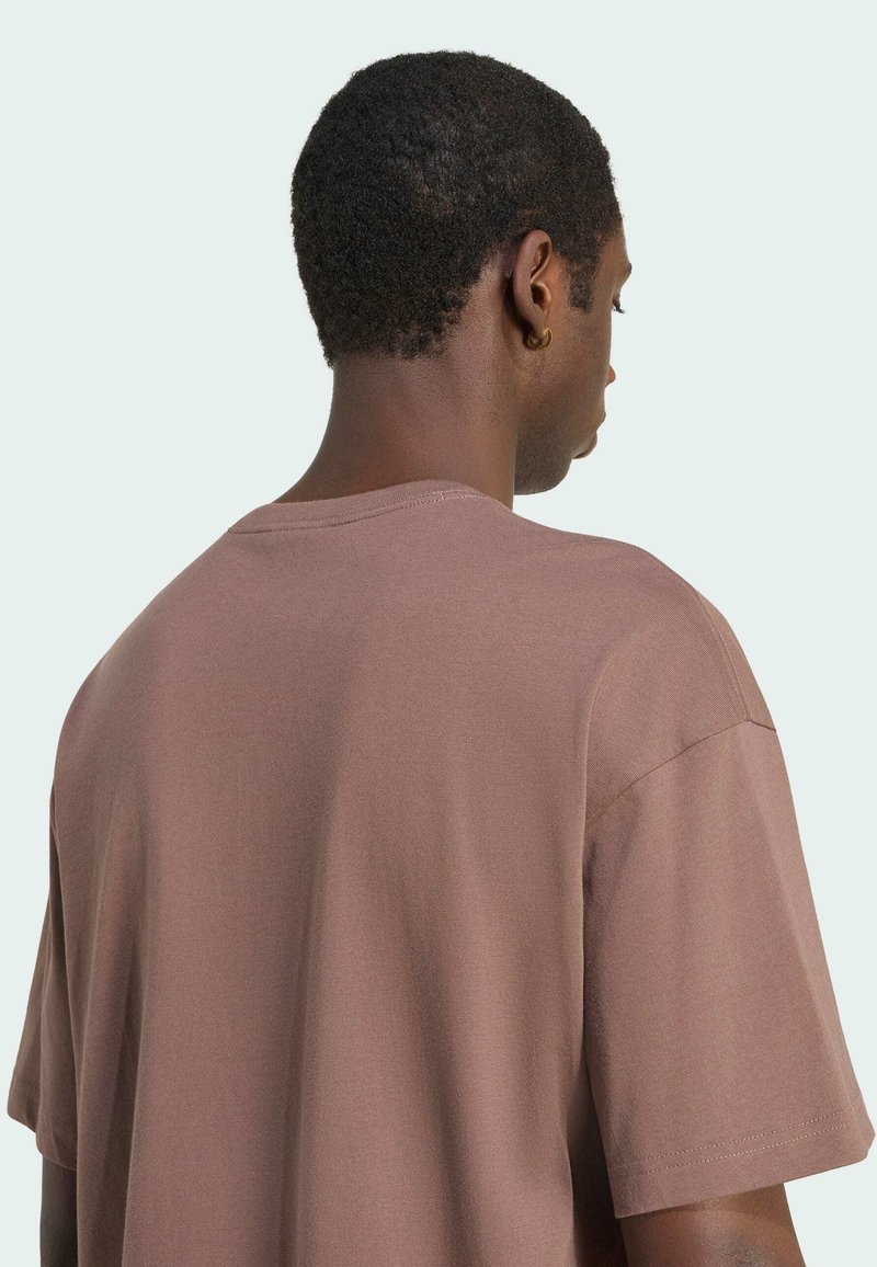 Homme aux cheveux courts et bouclés portant une chemise marron oversize à manches courtes, vu de dos et de profil sur un fond uni.