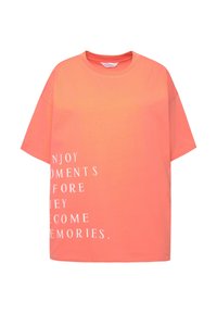 Camiseta de gran tamaño color coral hecha de algodón, con mangas cortas y cuello redondo. Texto impreso en blanco en el lado izquierdo.
