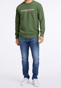 Felpa verde a maniche lunghe con "COPENHAGEN" e "LINDBERGH" stampati in bianco e arancione. Abbinata a jeans blu e sneakers bianche.