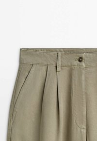 Pantalon beige taille haute avec plis devant, poches latérales, passants de ceinture et fermeture par bouton sur fond uni.