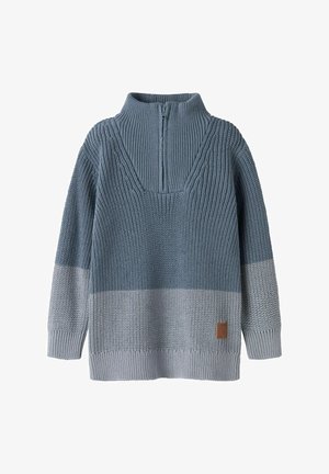 Pull tricoté bleu bicolore avec un col montant à fermeture éclair partielle et des manches longues, présentant un motif texturé et une petite pièce marron près de l'ourlet.