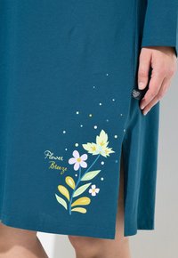 Turkusowa spódnica z bocznym rozcięciem, wzór kwiatowy w kolorach żółtym, różowym i zielonym, z napisem "Flower Breeze" blisko końca, ręka oparta obok rozcięcia.