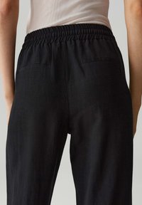 Zwarte broek van gestructureerde stof, met een elastische tailleband en twee achterzakken, gezien van de achterkant.