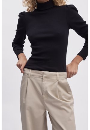 Osoba ubrana w czarną bluzkę z długim rękawem i bufkami na ramionach oraz beżowe plisowane spodnie z wysokim stanem, dłonie regulujące pas.