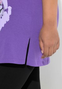 Fioletowy bawełniany t-shirt z bocznym rozcięciem, z grafiką w jasnych kolorach. Materiał wydaje się gładki, o casualowym kroju.