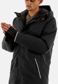 Chaqueta aislante negra con cuello alto, puños ajustables y detalles reflectantes. Cuenta con una cremallera y bolsillos laterales para almacenamiento.