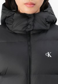 Gros plan d’une personne portant une doudoune noire avec une fermeture éclair devant, un col montant, des boutons-pression, et un logo blanc "cK" sur la poitrine.