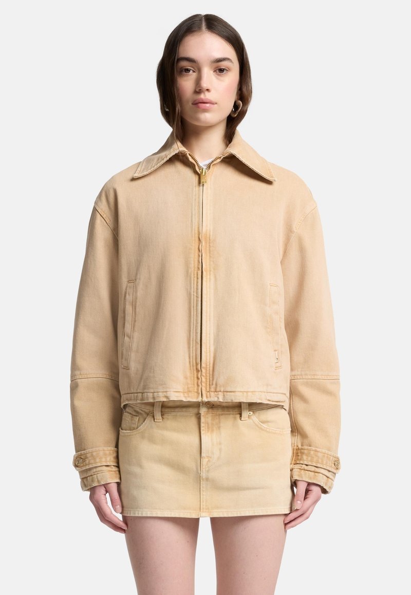 Beżowa kurtka dżinsowa z klasycznym kołnierzem, zamkiem błyskawicznym i opuszczonymi ramionami. Posiada przednie kieszenie i krótki krój.