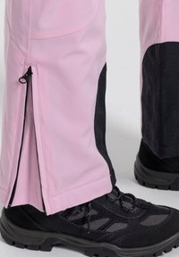 Ljust rosa vattentäta byxor med svarta detaljer, som har sidodragkedjor, flexibelt material och en smal design, kombinerade med svarta vandringsskor.