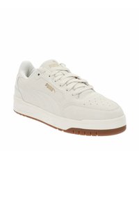 Zapatilla de ante blanco con punta redondeada, parte delantera con cordones y suela de goma marrón. Detalles de logotipo dorado y perforaciones en la punta para ventilación.