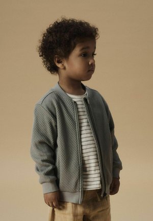 Veste bomber grise texturée avec des poignets et un col côtelés, portée sur une chemise blanche à rayures. L'enfant a les cheveux courts et bouclés et se tient de profil.