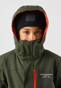 Grön vattentät jacka med orange innertyg, hög krage och logotyp. Kombinerad med en svart stickad mössa med läderpatch.