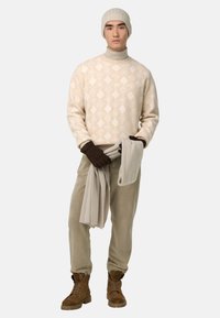 Pull crème en tricot avec motif géométrique, col roulé beige, gants marron, pantalon beige clair et bottes en daim marron, tenant une écharpe côtelée.