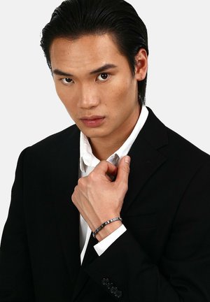 Silverfärgad armband med rektangulära länkar, prydd med ett geometriskt mönster, bärs på ett handled. Svart kostym med vit skjorta. Slät textur.