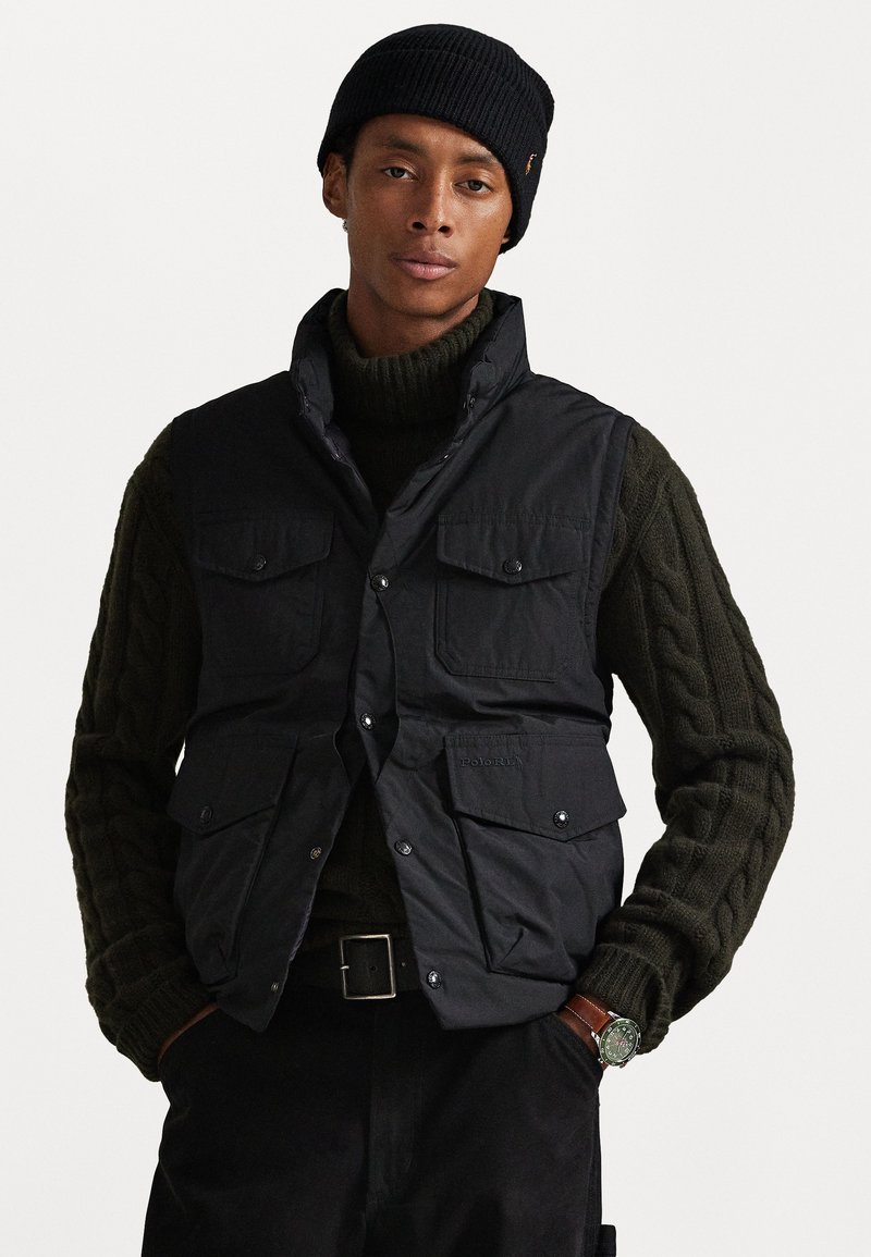 Black padded vest with four front pockets, zip closure, and snap buttons, layered over a dark green cable-knit sweater.