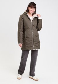 Manteau isolant vert olive avec fermeture éclair, capuche et doublure en polaire. Comprend deux poches latérales zippées et une coupe droite.