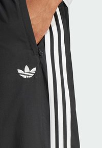 Svarta atletiska shorts med en vit Adidas-logotyp och tre vertikala vita ränder längs sidan. Slät textur, synlig ficka med dragkedja.