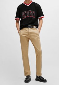 Svart kortärmad T-shirt med röd och vit kant som har "HUGO"-logotypen. Kombinerad med beige smala byxor och svarta skor med dekorativ hårdvara.