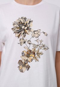 Hvit t-skjorte med blomstermønster med svarte konturer og aksenter i gult og oransje, som viser flere blomsterdesign og blader.