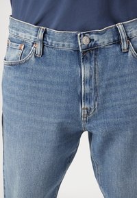 Μπλε τζιν παντελόνι με ίσια γραμμή, με φωτεινό πλύσιμο, χαλκές ραφές, ασημένιο κουμπί και παραδοσιακό σχεδιασμό πέντε τσεπών.