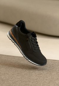 Baskets décontractées noires avec lacets et fermeture éclair sur le côté, semelle texturée et accents marron sur la semelle intermédiaire, posées sur un tapis beige dans un cadre intérieur neutre.