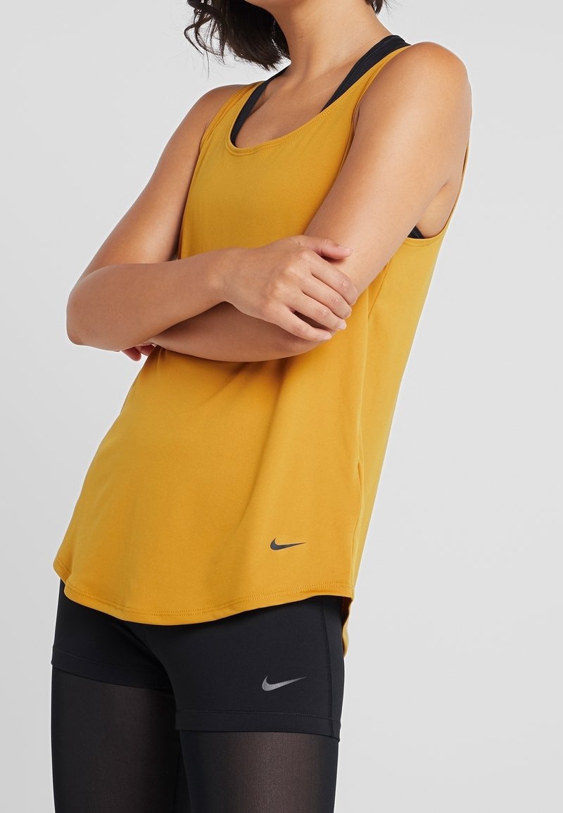 Femme portant un haut sans manches jaune moutarde et un short noir avec des logos Nike, bras croisés devant un fond uni.