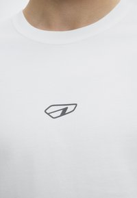Κλείσιμο σε ένα άτομο που φοράει λευκό t-shirt με στρογγυλή λαιμόκοψη και μικρό μαύρο γεωμετρικό λογότυπο στο στήθος.