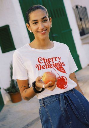 Nainen hymyilee ulkona ja pitää kädessään omenaa, pukeutuneena valkoiseen "Cherry Delicious" -t-paitaan ja korkeavyötäröisiin sinisiin farkkuihin, joissa on solmittu vyö.
