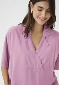 Lilac blūze ar V-veida dekoltē un īsām piedurknēm, izgatavota no mīksta auduma. Iezīmējas ar smalku teksturētu apdari un brīvu piegriezumu.