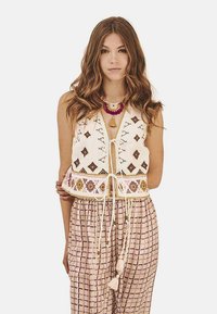 Chaleco de algodón bordado con patrones geométricos en marrón y rosa, que presenta un cierre con cordón. Combinado con pantalones de rayas y corte holgado.