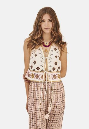 Chaleco de algodón bordado con patrones geométricos en marrón y rosa, que presenta un cierre con cordón. Combinado con pantalones de rayas y corte holgado.