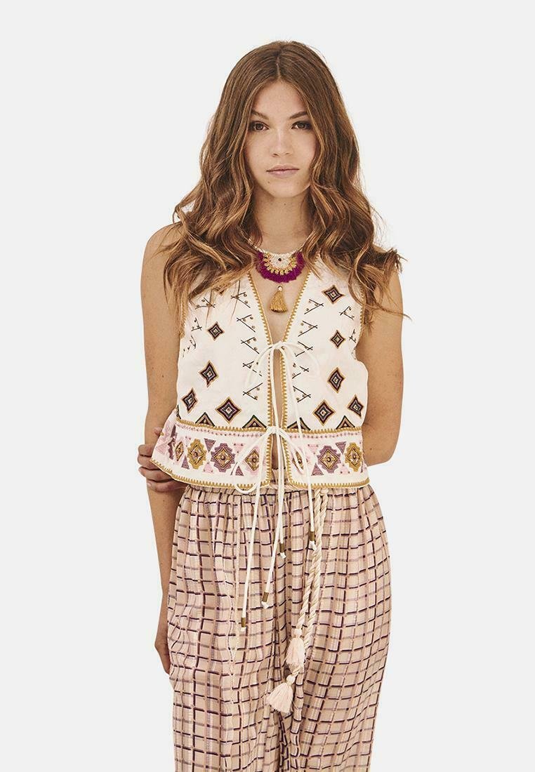 Chaleco de algodón bordado con patrones geométricos en marrón y rosa, que presenta un cierre con cordón. Combinado con pantalones de rayas y corte holgado.