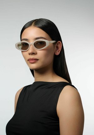 Vrouw met steil zwart haar, beige cat-eye zonnebril en een mouwloos zwart topje tegen een effen grijze achtergrond.