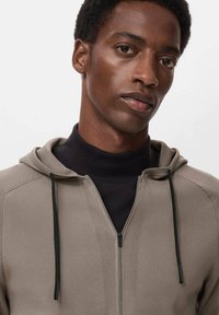 Lichtbeige rits-hoodie met een gestructureerde afwerking, zwarte trekkoordaccenten en een nauwsluitende coltrui eronder, tegen een neutrale achtergrond.