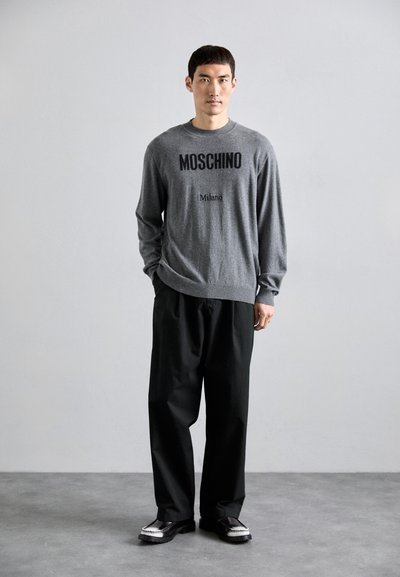 Pilkas megztinis su juodu užrašu "Moschino Milano", derinamas su laisvomis juodomis kelnėmis ir juodais bateliais su baltais akcentais. Paprastas, šiuolaikiškas dizainas.