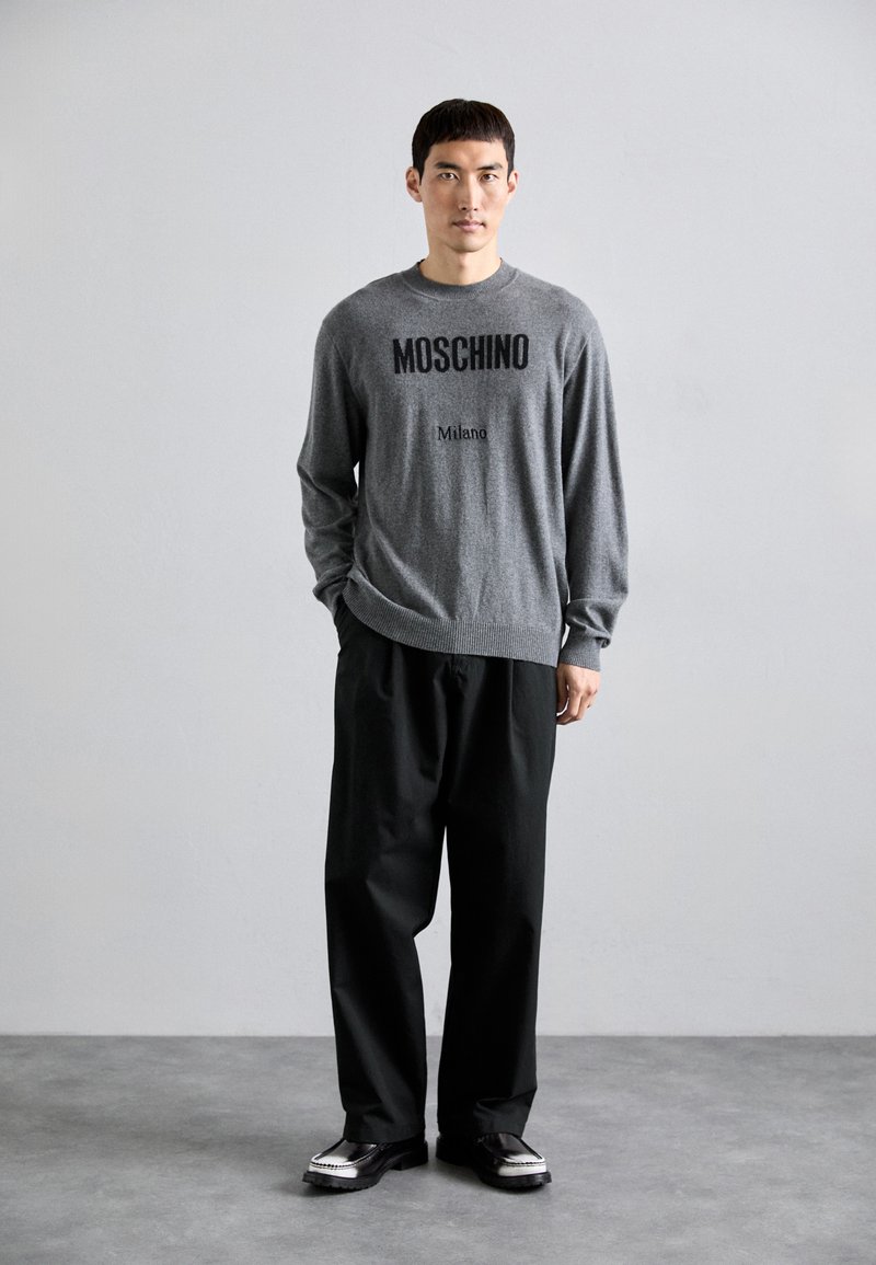 Grauer Pullover mit schwarzem "Moschino Milano"-Text, kombiniert mit lockeren schwarzen Hosen und schwarzen Schuhen mit weißen Akzenten. Schlichtes, modernes Design.