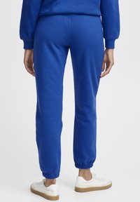 Blå sweatpants lavet af blødt stof, med elastiske manchetter og talje, parret med hvide sneaker og en glat tekstur.