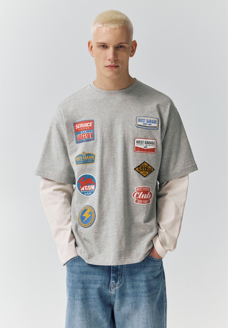 Giovane uomo con capelli biondi corti che indossa una camicia bianca a maniche lunghe a strati e una T-shirt grigia con toppe colorate a tema garage, jeans blu.