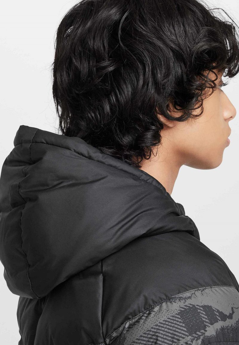 Chaqueta de plumas negra con capucha, textura suave y un acento gris con patrón en el hombro. La vista muestra el perfil lateral del modelo.
