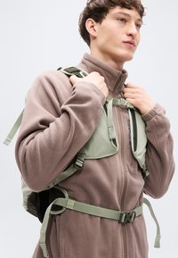 Ζακέτα από φλις σε απαλό καφέ χρώμα με ψηλό γιακά· φοράει πράσινο σακίδιο πλάτης σε στυλ ιμάντα με ρυθμιζόμενους ιμάντες και κούμπωμα με αγκράφα.