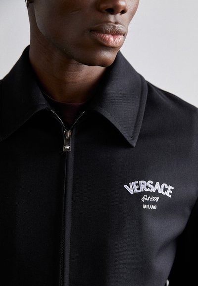 Sort jakke med krave, lynlås foran, og hvid broderet "VERSACE" logo. Glat stof med en skræddersyet pasform.