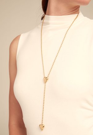 Collar largo en tono dorado que presenta dos colgantes en forma de corazón pulidos, conectados por una delicada cadena, llevado sobre un top blanco sin mangas.