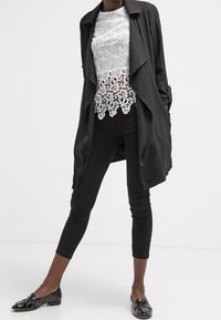 Cappotto trench nero con un design a strati, abbinato a un top in pizzo bianco e pantaloni neri aderenti, con eleganti mocassini neri lucidi.
