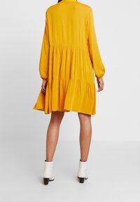 Gelbes, gestuftes Kleid mit langen Ärmeln, aus weichem Stoff gefertigt. Es hat einen runden Halsauschnitt und elastische Bündchen. Getragen mit weißen Knöchelstiefeln.